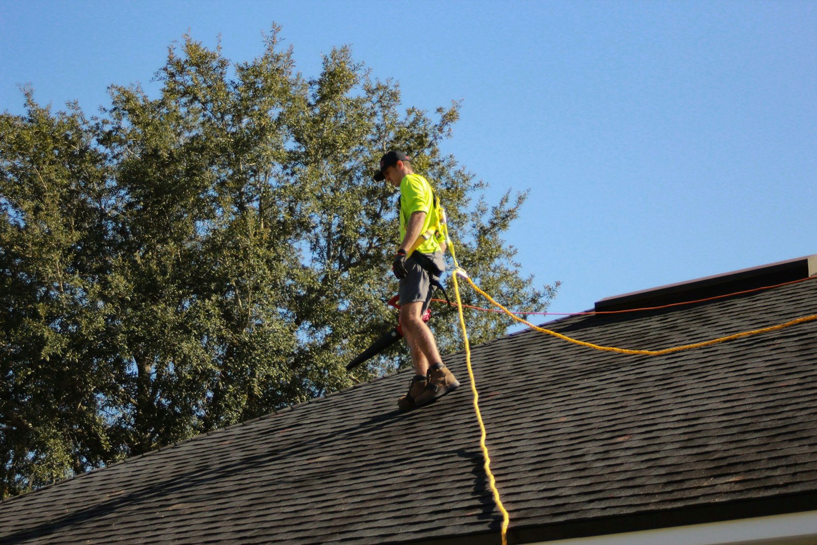 A roofer in a high-visibility vest and safety harness working on a shingle roof against a clear blue Southern Utah sky