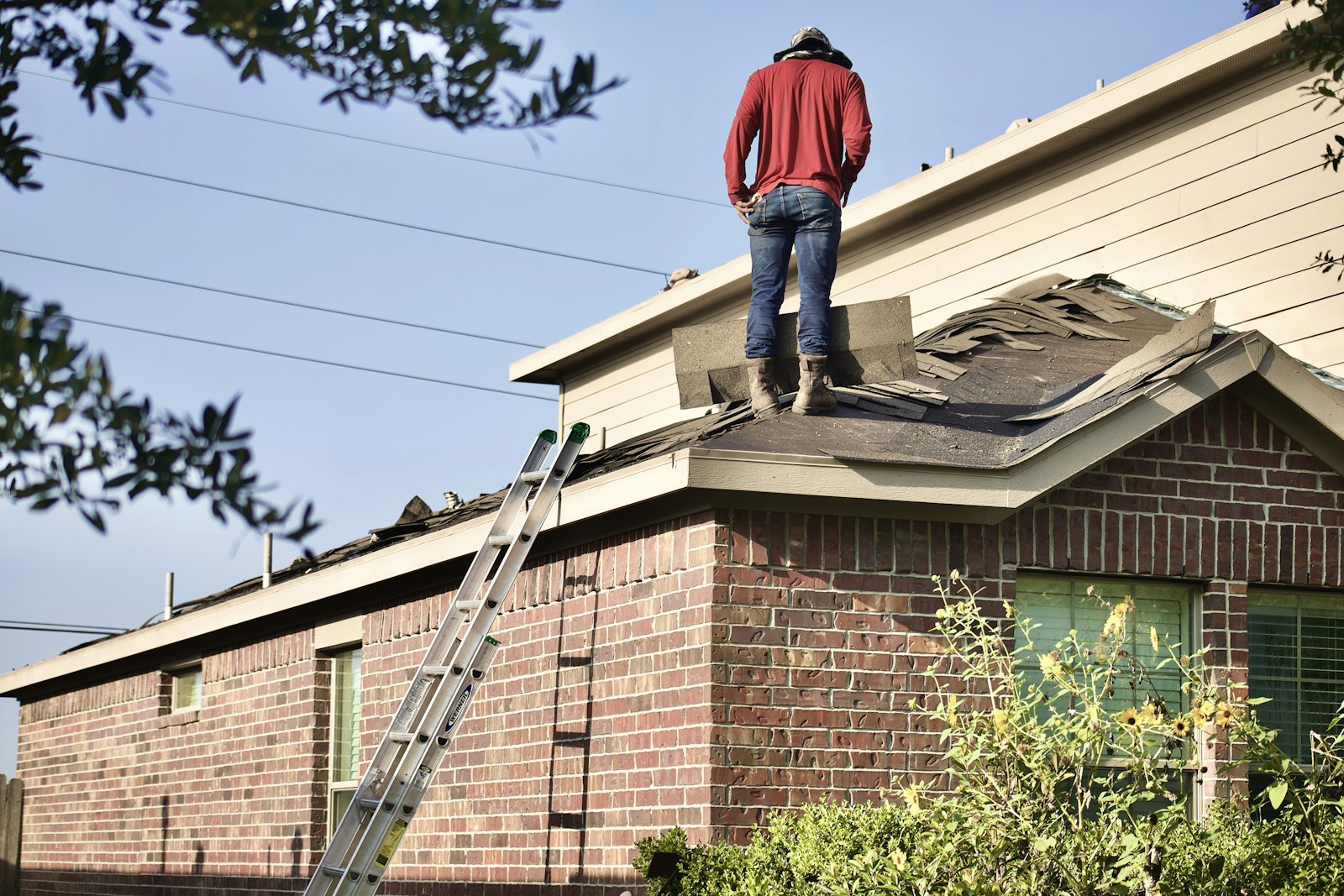 A roofer tearing off old shingles on a residential roof with a ladder propped against the brick exterior