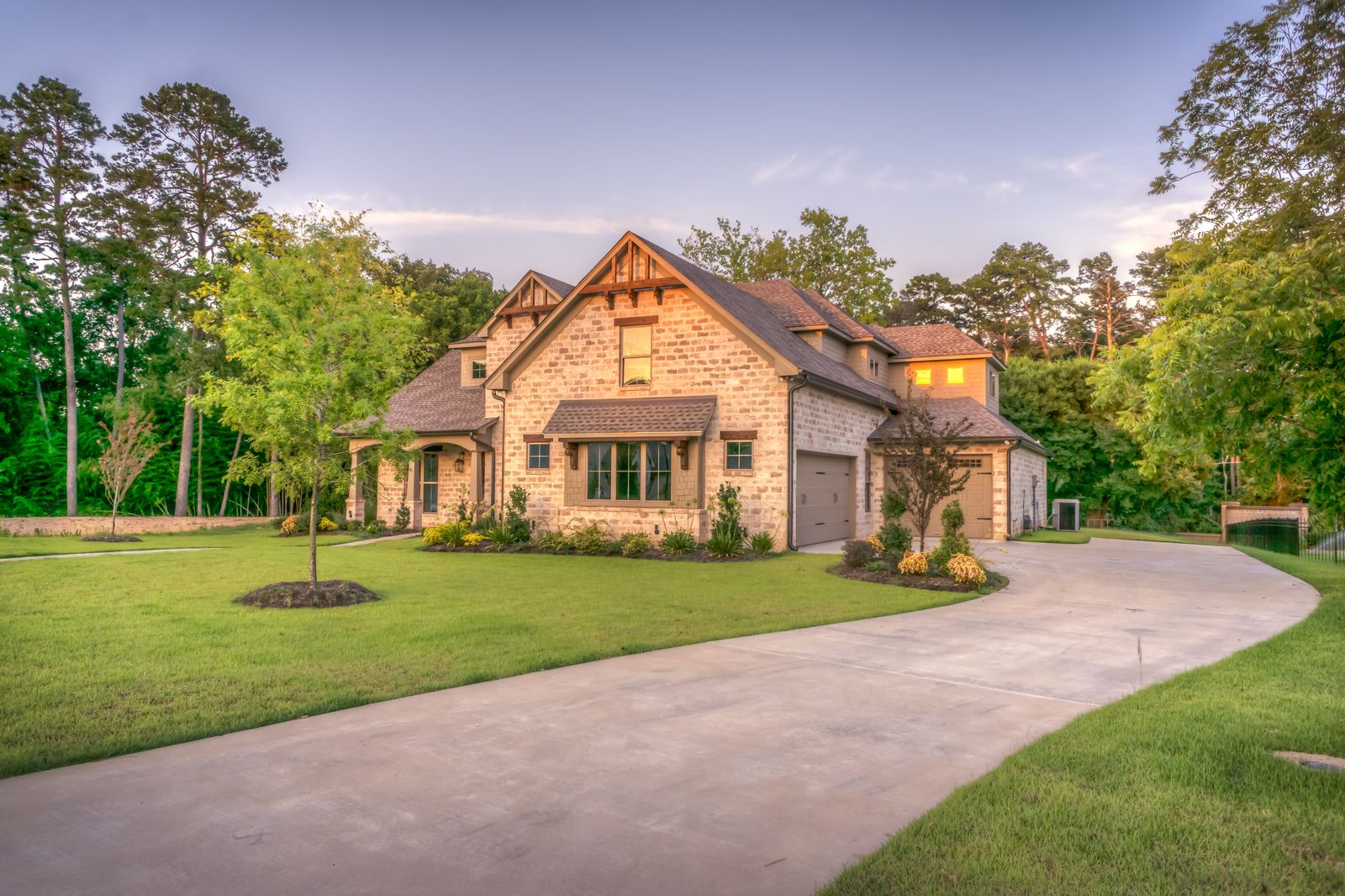 A large stone-clad home with a clean asphalt shingle roof glowing at golden hour, framed by trees and a wide driveway
